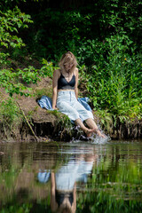 A girl with blond hair on the lake shore has her feet in the water and is swinging them creating a spray of water. Girl in a denim suit in nature