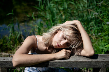 The girl with the blond hair on the lake shore rested her elbows and head on the bench. A thoughtful woman in a dress. Rest, relaxation, recuperation.