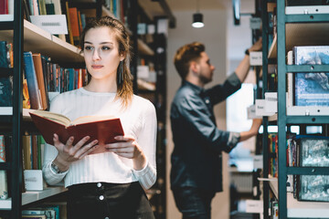 Serious female college student in casual wear pondering on literature standing near bookshelves in store,attractive young woman librarian concentrated on job holding book making order in library. - obrazy, fototapety, plakaty