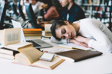 Young woman sleeping at desktop near books and literature in university campus overworked during making coursework project, smartphone with blank screen on working place, exhausted female student .