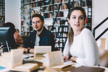 Portrait of skilled male student in eyewear learning and doing homework in university library using technology and wifi sitting next to female student, selective focus on smart young man at desktop