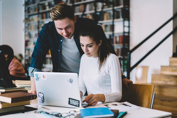 Young male and female colleagues watching tutorial together on laptop computer connected to wifi in college campus, skilled hipster guy helping girlfriend update application on netbook for learning