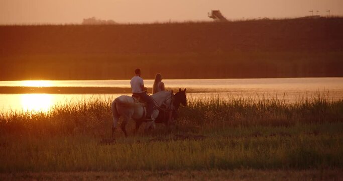 Caucasian couple ride horses by lake during golden hour. Medium wide shot panning handheld camera in slow motion. 