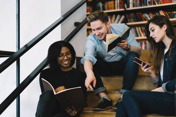 Cheerful male and female students joking and having fun while reading books in college library,...