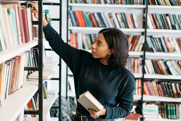 Young female african american librarian making order on bookshelves in college, dark skinned woman student picking literature for education checking information in books store, hipster girl get novels - obrazy, fototapety, plakaty