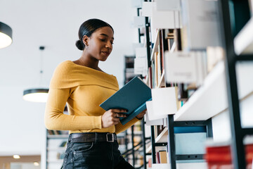 Pensive african american young woman attentively reading information from textbook preparing for seminar in library.Concentrated dark skinned student dressed in casual wear holding book in hands