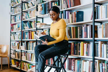 Concentrated african american young woman in eyeglasses for vision corrective reading literature book preparing for exam.Pensive dark skinned student searching information in textbook for coursework