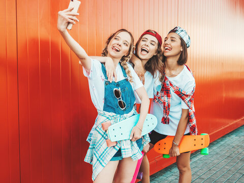 Three young smiling beautiful girls with colorful penny skateboards.Women in summer hipster clothes posing in the street near red wall.Positive models having fun and taking selfie self portrait photos