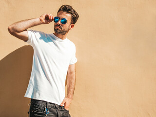 Portrait of handsome confident stylish hipster lambersexual model.Man dressed in white T-shirt. Fashion male posing in the street near wall in round sunglasses