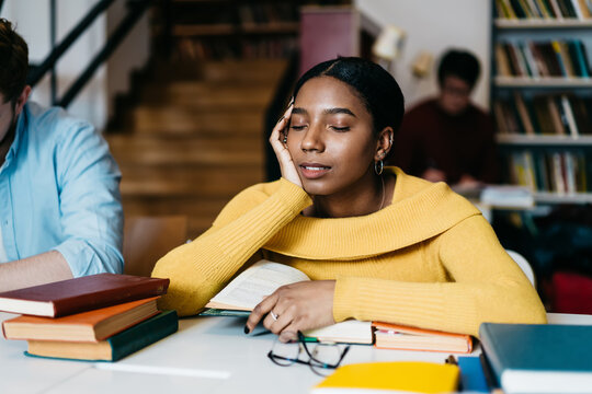 Tired african american student dressed in casual wear sleeping while sitting at desktop with many literature books for exam preparation in modern classroom during conducting boring studying lesson