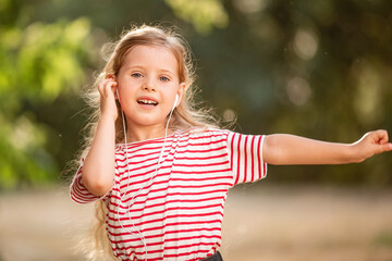 Happy little blonde girl listening to music with headphones, dancing and singing in nature in the Park.