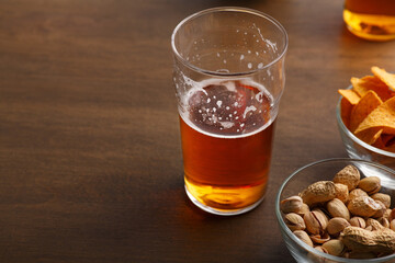 Half glass of ale in glass stands on table, near pistachios and nachos in bowls