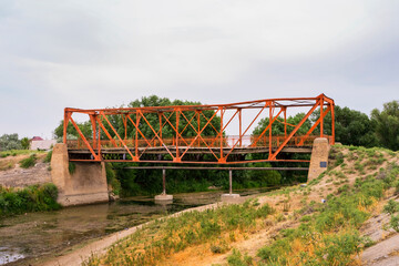 Ancient village bridge over the river. The metal frame of the Red Bridge. Wooden boards of the old bridge. Historical monument. Middle Asia. Kazakhstan. Country road. Green grass and sky