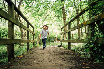 4-year-old brown boy running head-on across a bridge in a beautiful green forest with yellow sunlight
