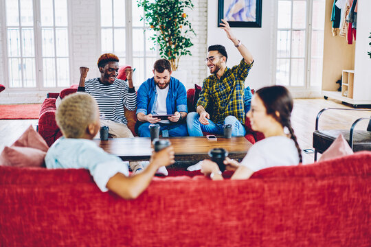 Excited multiracial guys raising hand up celebrating victory of male friend pass new level in video game browsed on tablet in coworking space, happy  men cheering for colleague playing online .
