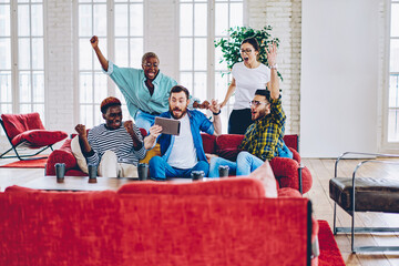 Overjoyed multiracial group of male and female fans cheering for soccer game watching on digital tablet together in loft interior, happy emotional hipsters expressing surprise with victory of contest.