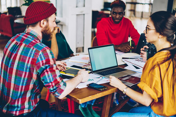 Young male and female students cooperating in coworking space using laptop computer with mock up screen, multiracial crew of skilled employees talking and discussing ideas during meeting table .