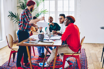 Skilled male employee presenting his strategy to multiracial crew of colleagues sitting together at meeting table, young creative team discussing ideas and opinions cooperating on project planning