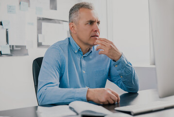 Fototapeta premium handsome gray-haired man sits in his office holding hand near face looking surprised while working on business project, work routine concept