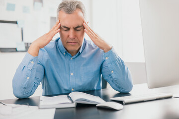 handsome tired gray-haired man sits in his office holding hands near temples looking exhausted while working on business project , stress at workplace concept