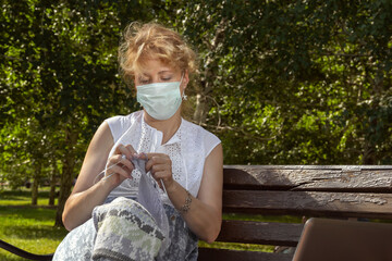 Woman in a medical mask on her face sits on a park bench and knits wool clothes on knitting needles. Hobbies in isolation