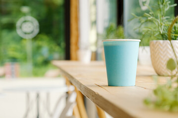 A blue paper cup with hot fresh coffee is standing on a wooden table in a cafe. The interior of the cafe with a Ripsalis cactus (twig) and other green plants. The view from the coffee shop.