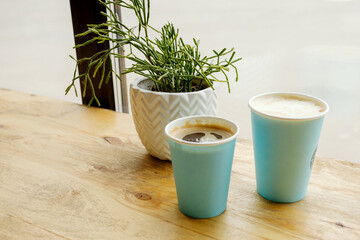 Blue paper glasses with hot fresh coffee stands on a wooden table in a cafe. The interior of the cafe with a Ripsalis cactus (twig) and other green plants. The view from the coffee shop.