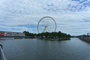 Montreal Grand Ferris Wheel in Old Port. Background is a blue cloudy sky. Foreground is trees and Saint Laurent River.