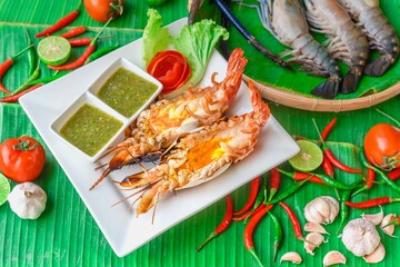 Grilled giant river prawn on white dish with spicy seafood sauce on green banana leaf background. Popular Thai food.