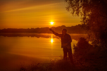 Morning on the lake dawn. A fisherman is standing on the shore next to fishing rods.