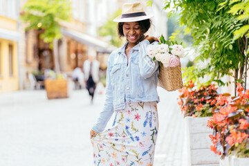 portrait of nigerian woman in straw sun hat, holding wicker bag with bouquet of peonies walking...