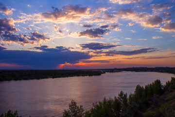Sunset over the river. The sky is purple and the sun is shining through the clouds. Twilight. Evening landscape with river and city in the background.