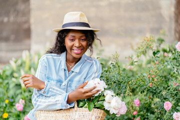 African woman in straw hat holding wicker bag with peonies, enjoying summer, walking on city street outdoor. Portrait of positive friendly nigerian girl, say Hi, sniffing flowers.