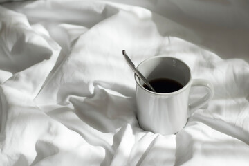 White coffee mug and spoon lay on the bed with the light from the window in the morning.