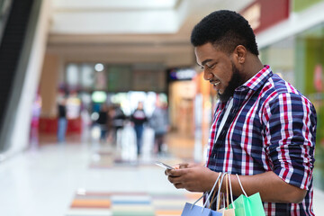 App With Discounts. Smiling Black Man Using Smartphone In Shopping Mall