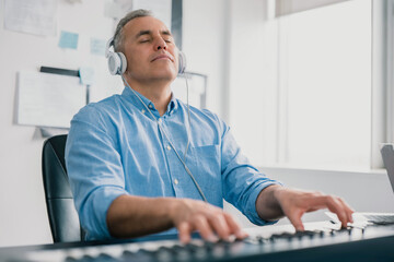 handsome gray-haired smiling man with headphones sits in music studio playing keyboard piano...