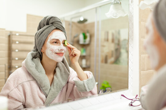 Beautiful young woman with towel wrapped around her head applying clay face mask at the bathroom