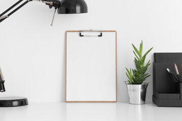 Wooden clipboard mockup wtih a aloe vera in a pot, lamp and workspace accessories on a white table.