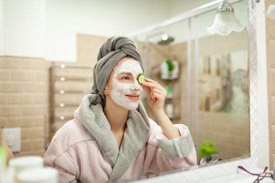 Beautiful young woman with towel wrapped around her head applying clay face mask at the bathroom