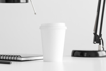 Coffee paper cup mockup with a lamp, notebook and a pen on a white table.
