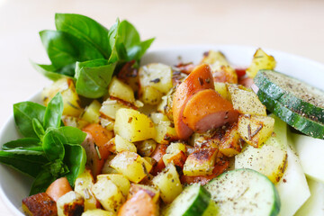 Saisonal und regionales Mittagessen, Bowl mit Bratkartoffeln mit Wiener Würstchen, gewürzt mit Rosmarin, Majoran, Pfeffer, Gurke,  Kohlrabi mit Basilikum als Beilage, deftiges kraftiges Mahlzeit