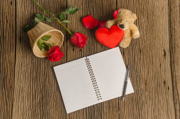 A photo of notepad on wood board with teddy bear holding heart and red rose