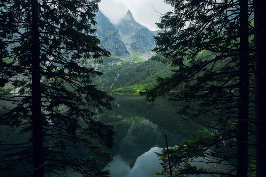 Beautiful view of foggy mountains cover by dark clouds and green forest with a reflection in a lake. Stony shore. Morskie Oko. Marine Eye. High Tatras, Zakopane