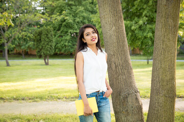 Young beautiful Indian girl standing in the park near a tree, holding a yellow notebook in her...