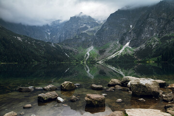 Beautiful view of foggy mountains cover by dark clouds and green forest with a reflection in a lake. Stony shore. Morskie Oko. Marine Eye. High Tatras, Zakopane