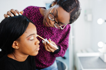 African American woman applying make-up by make-up artist at beauty saloon.