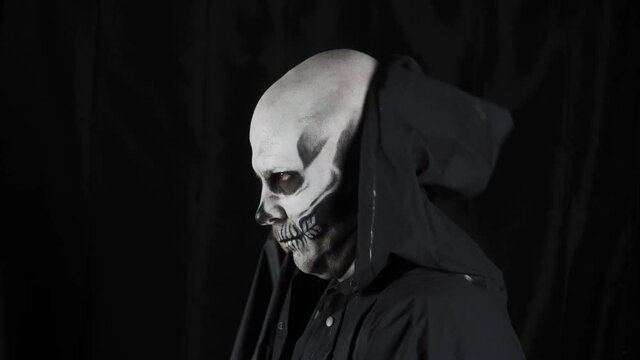 Man with make-up skeleton and black hood on a dark background. The skeleton takes off the hood and looks at the camera. Halloween or horror theme. High quality 4k video. High quality 4k footage