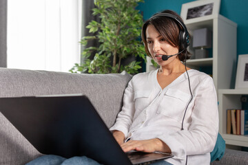 Beautiful woman in white shirt and blue jeans sitting comfortably on grey couch and using headset for video call on laptop. Concept of technology and remote work.