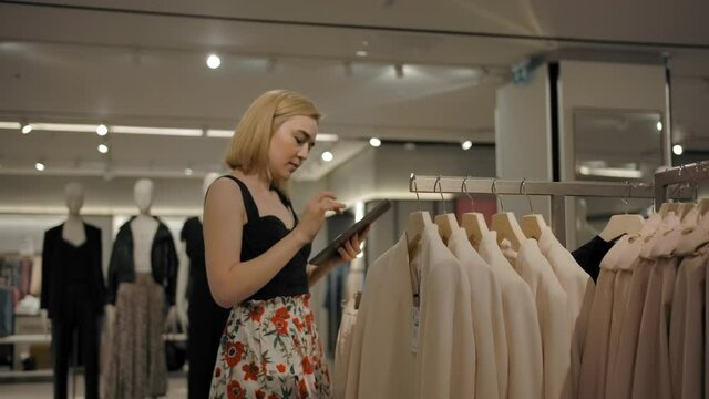 Young blond female examining clothes on rails and browsing tablet during weekend shopping in mall.