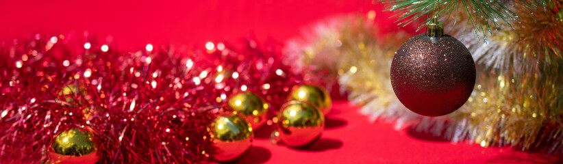 Christmas banner: a brown and gold Christmas ball weighs on a branch of a Christmas tree on a red background next to red tinsel and Golden balls. Side view, space for text.